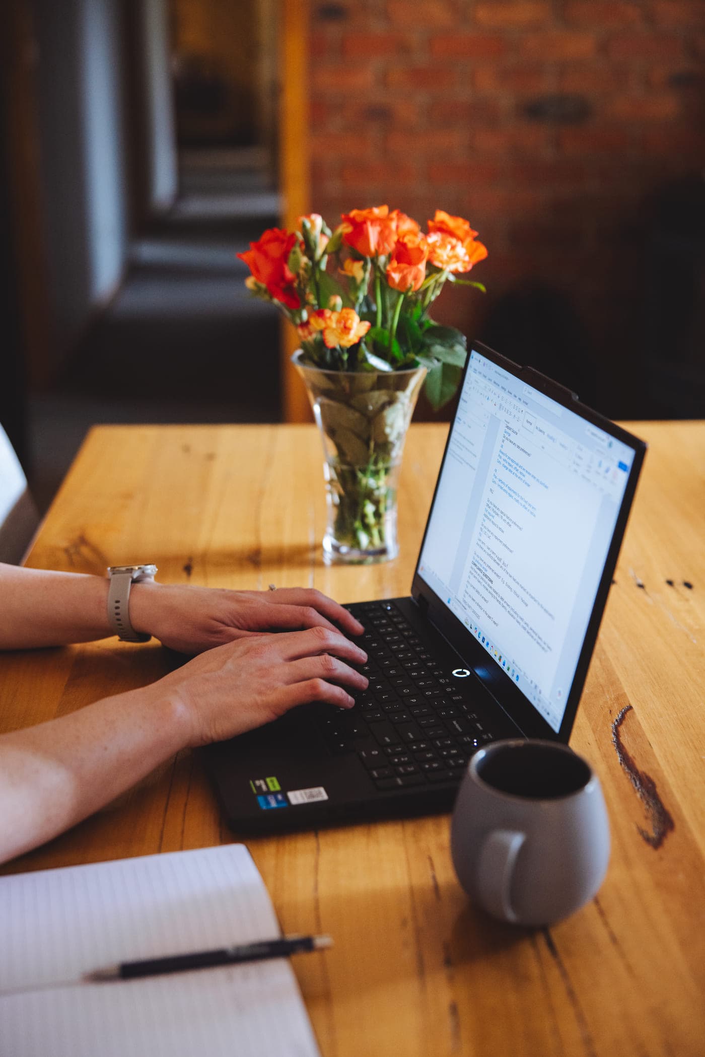 Alisha's hands on her laptop keyboard at the kitchen table, with a notebook, a coffee mug and orange roses beside her