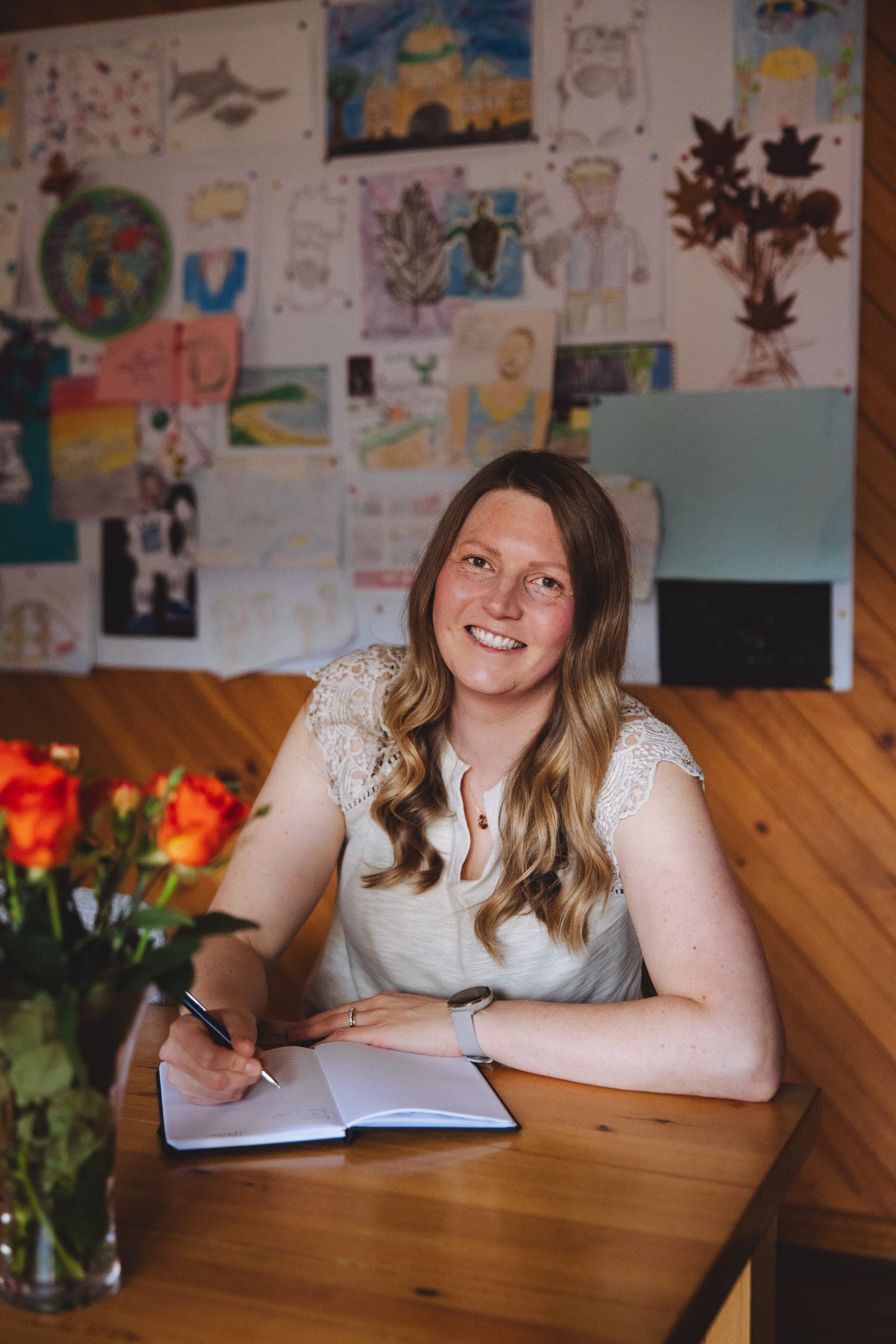Alisha Perry at her kitchen table, writing in a notebook beside her laptop and a vase of orange roses, with her children's drawings pinned on the wall behind her